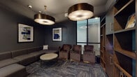 This single-floor amenity space combines upscale lounge design with an entertaining golf simulator experience. The lounge area is defined by deep charcoal and slate-toned walls that create a moody, sophisticated atmosphere, complemented by patterned carpet flooring in a herringbone-style layout. Seating is generous and thoughtfully arranged, featuring a large sectional sofa in warm grey upholstery alongside plush velvet accent chairs in rich brown tones, all centered around a round stone-topped coffee table with a metal wire base. Overhead, oversized drum pendant lights with bronze finishes serve as striking focal points, casting a warm, ambient glow throughout the lounge. Curated abstract artwork in bold blue and green hues adorns the dark walls, adding a contemporary artistic touch. Built-in wooden shelving provides both storage and display space, while glass-paneled doors connect the lounge to an adjoining corridor, maintaining an open and accessible flow. The golf simulator room is a dedicated, darkened space with matte black walls and a full-scale projection screen, complete with a synthetic turf hitting mat. Decorative circular canvas art adds personality to this entertainment zone. Natural light filters into the lounge through large windows, offering a balanced contrast to the intentionally dramatic interior palette.