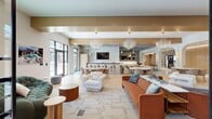 This generously sized single-floor amenity space is thoughtfully designed to serve as a premier social hub. The open-concept main lounge is anchored by striking globe pendant chandeliers and features light hardwood flooring throughout, creating a warm and inviting atmosphere. Curated seating arrangements — including terracotta sofas, sculptural lounge chairs, and round dining tables with upholstered chairs — offer flexible spaces for both casual gatherings and more formal socializing. Bold abstract wall art adds a contemporary artistic touch.

A full wet bar and catering kitchen serve as the heart of the entertaining area, complete with a dark stone countertop island with bar seating, white textured tile backsplash, a wall-mounted TV, open shelving with curated décor, and light wood cabinetry with brass hardware. The adjacent full catering kitchen features herringbone tile flooring, ample counter space, and direct views to the outdoor pool area through large black-framed windows and glass doors.

A separate media lounge provides a more intimate retreat, highlighted by wood-paneled ceiling accents, a built-in TV console with warm wood cabinetry, bold geometric rugs, and distinctive curated artwork. Throughout the space, large floor-to-ceiling windows and glass entry doors flood the interiors with natural light and offer views of the outdoor amenities.