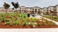 This residential community showcases a Mediterranean-inspired architectural design, characterized by a neutral stucco exterior, tiled rooflines, and warm accent details. The multi-story buildings are arranged around a central, generously sized communal courtyard that serves as the heart of the community.

The outdoor common area is thoughtfully designed for both relaxation and social interaction. A well-maintained lawn provides open green space, while a paved patio area accommodates a picnic table and a barbecue grill, making it well-suited for gatherings and outdoor dining. Decorative stepping stone pathways wind through richly mulched garden beds, which are planted with a diverse mix of ornamental grasses, flowering shrubs, and young trees — lending the space a vibrant, park-like atmosphere.

Wooden trellis structures are positioned throughout the landscaped areas, adding architectural interest and supporting additional plantings as they mature. Private balconies and staircases are visible on the building façades, offering residents personal outdoor space in addition to the shared amenities below. The overall design reflects a strong emphasis on community living, combining attractive landscaping with practical outdoor features in a cohesive and inviting setting.