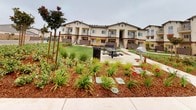 This residential community showcases a Mediterranean-inspired architectural design, characterized by a neutral stucco exterior, tiled rooflines, and warm accent details. The multi-story buildings are arranged around a central, generously sized communal courtyard that serves as the heart of the community.

The outdoor common area is thoughtfully designed for both relaxation and social interaction. A well-maintained lawn provides open green space, while a paved patio area accommodates a picnic table and a barbecue grill, making it well-suited for gatherings and outdoor dining. Decorative stepping stone pathways wind through richly mulched garden beds, which are planted with a diverse mix of ornamental grasses, flowering shrubs, and young trees — lending the space a vibrant, park-like atmosphere.

Wooden trellis structures are positioned throughout the landscaped areas, adding architectural interest and supporting additional plantings as they mature. Private balconies and staircases are visible on the building façades, offering residents personal outdoor space in addition to the shared amenities below. The overall design reflects a strong emphasis on community living, combining attractive landscaping with practical outdoor features in a cohesive and inviting setting.