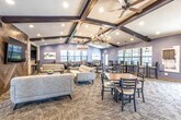 This single-story clubhouse is thoughtfully designed to accommodate a variety of social and recreational activities, offering multiple distinct zones within one cohesive space. The main lounge area serves as a central gathering point, featuring plush gray sofas arranged around a wooden coffee table beneath an eye-catching herringbone wood-paneled accent wall with a wall-mounted television. Dark exposed beams crisscross the coffered ceiling overhead, complemented by rustic chandelier lighting that adds warmth to the space.

Adjacent to the lounge, a spacious dining and café-style seating area offers an assortment of high-top and standard tables with black ladder-back chairs, illuminated by pendant lighting and lined with large windows that bring in generous natural light. A well-equipped full kitchen features dark espresso cabinetry, granite countertops, stainless steel appliances, and a breakfast bar with seating.

The recreation area includes a regulation-size billiards table positioned beneath a statement chandelier, alongside a fireplace with a decorative stone surround and a large flat-screen television. A quiet reading nook with a leather chair and oversized windows provides a more intimate retreat. Throughout, blue-gray walls, mixed flooring of hardwood and carpet, and consistent upscale fixtures create a polished, inviting ambiance.