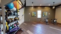 Located at 22 Campbell Scenic Dr in Hattiesburg, Mississippi, this single-floor fitness studio is thoughtfully designed to support a wide range of workout activities. The space is anchored by durable wood-look flooring with contrasting inlay accents, lending a clean and polished aesthetic throughout.

The main workout area is generously sized and open, allowing for flexible use across multiple fitness disciplines. A well-stocked vertical equipment rack holds an array of medicine balls, stability balls, kettlebells, resistance bands, battle ropes, and exercise mats — providing everything needed for functional and strength training. TRX suspension straps are mounted overhead for bodyweight training.

A dedicated cycling area houses multiple stationary spin bikes, positioned beneath natural light from multiple windows. Framed landscape artwork adorns the walls, contributing a calming visual element to the training environment. Ceiling-mounted pendant fans and recessed lighting ensure the space remains well-lit and comfortable during all types of workouts.

A wall-mounted flat-screen television is installed near the entrance, ideal for streaming classes or instructional content. Dark wood crown molding adds an elevated, refined finish to the room's upper perimeter. The layout flows naturally between zones, making this a highly functional and well-appointed fitness facility.