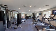 This single-floor fitness center offers a spacious, thoughtfully organized workout environment designed to support a full range of exercise routines. The interior is finished with neutral-toned walls, a textured ceiling with recessed lighting, and durable blue-toned carpet flooring that extends throughout the space.

The cardio zone is well-appointed, featuring multiple treadmills, elliptical trainers, and stationary bikes, including recumbent models. Wall-mounted flat-screen televisions are positioned throughout the room, providing entertainment during workouts. A dedicated corner area accommodates additional cardio machines alongside a window offering outdoor views.

The strength training section includes a multi-station cable machine, a robust selection of free weights with a full dumbbell rack, and multiple adjustable benches to support a variety of exercises. A large wall mirror spans one side of the room, visually expanding the space and serving as a practical training aid.

Natural light filters in through several windows along the perimeter walls, creating a bright and open atmosphere. An emergency exit door and clearly posted fitness center rules reflect attention to safety. The overall layout is practical and efficient, offering users ample space to move between equipment comfortably.