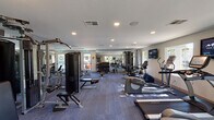 This single-floor fitness center offers a spacious, thoughtfully organized workout environment designed to support a full range of exercise routines. The interior is finished with neutral-toned walls, a textured ceiling with recessed lighting, and durable blue-toned carpet flooring that extends throughout the space.

The cardio zone is well-appointed, featuring multiple treadmills, elliptical trainers, and stationary bikes, including recumbent models. Wall-mounted flat-screen televisions are positioned throughout the room, providing entertainment during workouts. A dedicated corner area accommodates additional cardio machines alongside a window offering outdoor views.

The strength training section includes a multi-station cable machine, a robust selection of free weights with a full dumbbell rack, and multiple adjustable benches to support a variety of exercises. A large wall mirror spans one side of the room, visually expanding the space and serving as a practical training aid.

Natural light filters in through several windows along the perimeter walls, creating a bright and open atmosphere. An emergency exit door and clearly posted fitness center rules reflect attention to safety. The overall layout is practical and efficient, offering users ample space to move between equipment comfortably.