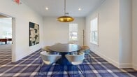 This residence lounge in Saugus, Massachusetts is a thoughtfully designed communal space spread across a single floor, offering a welcoming and sophisticated environment for residents. The layout encompasses a dedicated dining room and two distinct living areas, each with its own character and purpose.

The dining room is anchored by a large round dark-topped table surrounded by plush curved chairs, all set atop a bold blue plaid carpet. A striking gold pendant light hangs overhead, while large windows invite natural light and views of the outdoor surroundings. Abstract wall art adds a contemporary artistic touch.

The living areas offer versatile settings for relaxation and socializing. One space features a wall-mounted flat-screen television set against a warm terracotta accent wall, with decorative accents on a sleek console table below. The second living area showcases bold geometric murals in rust and cream tones, with modular gray banquette seating, bistro tables, and a mustard ottoman creating an inviting lounge atmosphere.

Throughout the lounge, a cohesive plaid carpet in blue and purple tones ties the spaces together, while large windows frame outdoor views and flood the interior with natural light. Potted greenery, curated artwork, and recessed lighting complete the polished, modern aesthetic of this vibrant communal retreat.
