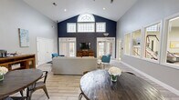 Located at Campus Pointe in Charleston, Illinois, this single-floor community space offers a generously proportioned open-plan layout combining a living room and dining area under one expansive roof. The most striking architectural feature is the dramatic vaulted ceiling, which rises to a considerable height and is anchored by a bold navy blue accent wall. A large arched transom window crowns this wall, flanked by a row of clerestory windows that flood the interior with natural light throughout the day. French doors along the same wall provide direct access to an outdoor patio area, further enhancing the sense of openness.

The living area is furnished with a neutral sectional sofa, vibrant teal accent chairs, and a tufted olive green ottoman arranged around a wood media console with a wall-mounted flat-screen television. Light wood-look tile flooring runs continuously throughout, lending cohesion to the entire space. The dining zone features multiple round dark wood tables paired with industrial-style metal chairs, complemented by decorative wall art in blue and gold tones. Interior windows connect this area to an adjacent room, possibly a fitness or business center, adding visual depth. The overall ambiance is modern, bright, and welcoming.