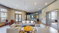 This single-floor community clubhouse offers a well-designed open-concept layout with distinct functional zones that flow seamlessly into one another. The main lounge area features warm hardwood-style flooring, recessed lighting, and generous seating arrangements including neutral-toned sofas and armchairs centered around a round wooden coffee table atop a patterned area rug. Large multi-pane windows and double French doors flood the space with natural light while providing views of an outdoor patio and pool area. A dedicated dining zone near the windows includes high-top tables and chairs, ideal for casual gatherings. The kitchenette bar is a standout feature, boasting a dark granite countertop, metallic subway tile backsplash, pendant lighting, and bar stools with a rustic reclaimed-wood base. A coffee station and sink complete the functional bar setup. An "Indiana"-themed sign adds regional character to the bold blue accent wall. The entry area includes a wall-mounted television and a striking floor-to-ceiling stacked stone fireplace, adding warmth and texture. A hallway connects to additional rooms beyond the main space, and the overall ambiance is modern yet inviting, balancing communal functionality with comfortable design.