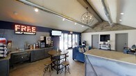 This single-floor space combines industrial and modern design elements to create a welcoming and functional communal lounge. The vaulted ceiling draws the eye upward, where exposed metal ductwork and a striking orb chandelier complement a track lighting system, together providing both ambient and task illumination. Stone-clad accent walls add texture and warmth, contrasting beautifully against the bold navy blue feature wall that anchors the space.

A self-serve coffee and beverage station lines one wall, equipped with coffee machines and a selection of refreshments, making it an ideal hub for socializing or quick refreshment breaks. A rustic round high-top table with industrial-style bar stools provides casual seating nearby. A plush navy sofa with decorative pillows offers a more relaxed lounge area, positioned to take advantage of the natural light that pours in through large glass patio doors. These doors open to an outdoor area, seamlessly blending interior and exterior spaces.

A granite-topped counter on one side adds a practical workspace or serving surface, while the polished concrete floors reinforce the space's urban, industrial aesthetic. The overall ambiance is modern yet inviting — a well-appointed gathering space designed for comfort and community.