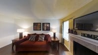 This single-floor space offers a warm and inviting atmosphere, thoughtfully designed with a blend of classic and comfortable elements. The living area serves as the heart of the home, showcasing rich hardwood flooring that anchors the room with a sense of warmth and character. A traditional fireplace with a clean white mantle serves as a natural focal point, adding both charm and functionality to the space. The walls feature a neutral-to-warm color palette, with soft white upper walls transitioning to a muted golden-yellow accent, lending the room depth and a cozy feel. A textured ceiling adds subtle architectural interest overhead. Natural light filters into the living area through a window dressed with light curtains, complementing the ambient lighting provided by tasteful floor and table lamps. The space is well-proportioned, offering a comfortable and practical layout suitable for relaxed everyday living. Decorative framed artwork on the walls and carefully placed accessories on the mantle contribute to the overall polished yet homey aesthetic. The combination of warm tones, classic architectural details, and thoughtful design makes this a welcoming and livable space.
