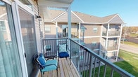 This single-floor unit offers a thoughtfully arranged open layout that connects the main living spaces seamlessly. The living room is bright and airy, anchored by light wood-style flooring and a large sliding glass door that fills the room with natural light and provides direct access to the private balcony. The balcony features wood decking, metal railing, and room for outdoor seating, overlooking the surrounding green landscaping and neighboring buildings.

The kitchen is efficiently designed with rich dark wood cabinetry extending to the upper walls, stainless steel appliances including a refrigerator, range, and built-in microwave, complemented by a breakfast bar that connects to the main living area. Recessed lighting and light countertops round out the functional workspace.

Adjacent to the kitchen, a compact dining area accommodates a round table and chairs, accented by decorative wall art that adds character to the space. Light wood-style flooring continues throughout the main areas, while carpeting transitions into the bedroom, adding warmth and comfort.

The unit's neutral wall tones provide a versatile backdrop throughout, and the open sightlines between rooms create a cohesive, comfortable living environment overall.