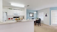 This single-story unit at Academy Circle in Kissimmee, Florida offers a thoughtfully arranged layout with distinct living, dining, and sleeping areas. The kitchen is designed as a galley-style space with white cabinetry, light countertops, wood-look flooring, and a full suite of white appliances including a refrigerator, range, microwave, and dishwasher. A peninsula with a light countertop extends into the adjoining area, creating a natural transition between the kitchen and the rest of the living space.

The dining area is defined by a soft blue accent wall, pendant lighting overhead, and a window with blinds that allows natural light to filter in. It comfortably accommodates a round dining table with seating for four. Adjacent to the dining space, the living room continues the blue color palette and features carpet flooring, a ceiling fan, and multiple windows that bring in additional natural light.

The bedroom offers generous proportions with carpet flooring, multiple windows with blinds, and a ceiling fan. A closet with sliding doors provides practical storage. The bathroom includes tile flooring, a single vanity with white cabinetry, and a shower-tub combination. A secondary room with carpet and closet space can serve as an additional bedroom or home office.