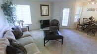 This single-floor residence centers around a welcoming living room designed for both comfort and functionality. The space is anchored by a tile-surround fireplace, which serves as a natural focal point and adds a classic, cozy character to the room. A decorative mirror with an ornate frame hangs above the mantle, enhancing the sense of depth and elegance in the space.

Plush wall-to-wall carpeting runs throughout the living area, contributing to a warm and inviting atmosphere underfoot. Natural light fills the room generously, streaming in through multiple windows fitted with white horizontal blinds, as well as a glass-paneled exterior door that offers direct access to the outdoors. This combination of windows and door ensures the space remains bright throughout the day while providing convenient outdoor connectivity.

The layout accommodates a comfortable seating arrangement, with ample room for a sofa, accent chair, and a tufted leather ottoman that doubles as a coffee table. The neutral wall tones complement the soft carpeting, creating a versatile backdrop suitable for a variety of décor styles. Overall, the living area presents a practical and pleasant single-level living environment.