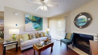 This single-story residence offers a well-organized layout visible through its open floor plan. The living room serves as the heart of the home, showcasing rich hardwood-style flooring, a textured ceiling with a ceiling fan, and a fireplace framed by a dark surround. A large sliding glass door fitted with vertical blinds fills the space with natural light and provides direct access to an outdoor patio area with views of surrounding greenery. Bold, colorful wall art adds a vibrant focal point to the room's neutral palette.

The galley-style kitchen features warm wood cabinetry, white appliances including a range, microwave, and refrigerator, and hardwood-style flooring that continues from the main living area. A pass-through connects the kitchen to the dining and living spaces, promoting an open feel. In-unit laundry access is visible at the rear of the kitchen corridor.

The bedroom is comfortably sized, finished with plush carpet, a ceiling fan, and a window dressed with patterned drapes and shuttered blinds that allow for natural light control. The bathroom presents a vanity with wood cabinetry, a mirrored wall, and a tub-shower combination, all complemented by textured walls and hardwood-style flooring.