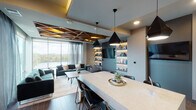 Located along The Boardwalk Trail in Austin, TX, this sophisticated single-floor space showcases a thoughtfully designed open-plan layout where the kitchen, dining, and living areas flow seamlessly into one another. Floor-to-ceiling windows along the living area's exterior wall frame sweeping waterfront views of what appears to be Lady Bird Lake, flooding the interior with natural light and creating a strong visual connection to the outdoors.

A striking geometric wood-panel ceiling detail anchors the living area, adding warmth and architectural dimension. The living space features plush emerald-green velvet seating and sheer layered drapery that complement the scenic backdrop. Built-in illuminated shelving lines one wall, providing both display space and ambient lighting.

The kitchen and dining area center around a generous marble-topped island with seating for multiple guests, illuminated by a trio of angular black pendant lights. Dark cabinetry pairs with a glossy white elongated hexagonal tile backsplash and stainless steel appliances, including a built-in beverage cooler. Wood-look tile flooring runs throughout, tying the spaces together cohesively. The overall ambiance is contemporary and refined, with a palette of warm neutrals, deep jewel tones, and metallic accents.
