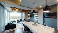 Located along The Boardwalk Trail in Austin, TX, this sophisticated single-floor space showcases a thoughtfully designed open-plan layout where the kitchen, dining, and living areas flow seamlessly into one another. Floor-to-ceiling windows along the living area's exterior wall frame sweeping waterfront views of what appears to be Lady Bird Lake, flooding the interior with natural light and creating a strong visual connection to the outdoors.

A striking geometric wood-panel ceiling detail anchors the living area, adding warmth and architectural dimension. The living space features plush emerald-green velvet seating and sheer layered drapery that complement the scenic backdrop. Built-in illuminated shelving lines one wall, providing both display space and ambient lighting.

The kitchen and dining area center around a generous marble-topped island with seating for multiple guests, illuminated by a trio of angular black pendant lights. Dark cabinetry pairs with a glossy white elongated hexagonal tile backsplash and stainless steel appliances, including a built-in beverage cooler. Wood-look tile flooring runs throughout, tying the spaces together cohesively. The overall ambiance is contemporary and refined, with a palette of warm neutrals, deep jewel tones, and metallic accents.