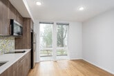This single-floor unit showcases a clean, modern design highlighted by warm hardwood flooring that runs throughout the main living areas. The open layout connects the living space to a well-appointed kitchen, which features dark wood cabinetry with sleek bar-pull hardware, white quartz countertops, stainless steel appliances including a refrigerator and built-in microwave, and a striking mosaic tile backsplash in muted tones of yellow, gray, and white that serves as a focal point of the space.

Large floor-to-ceiling windows and a glass sliding door in the living area flood the space with natural light while providing views of the surrounding greenery. The neutral wall palette and recessed lighting contribute to the bright, airy ambiance throughout.

The bathroom is finished with large-format tile flooring, a dark wood vanity with white countertop, an undermount sink, and a well-placed mirror with overhead lighting. Clean lines and consistent hardware finishes carry the contemporary aesthetic from room to room.

Additional features include a separate bedroom with its own large windows and access to an outdoor balcony, as well as a dedicated entryway with interior doors that provide clear separation between areas of the unit.