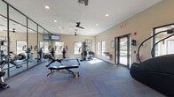 This fitness center is designed to provide a comprehensive workout environment within a single, open-plan space. The room is finished with durable, multi-toned blue carpet tiles laid in a herringbone pattern, neutral beige walls, and recessed lighting complemented by multiple ceiling fans that promote air circulation throughout. A defining architectural feature is the floor-to-ceiling mirror wall framed in dark metal, which visually expands the space while allowing users to monitor their form during exercise.

Natural light fills the room through numerous large windows positioned along multiple walls, offering views of the surrounding outdoor greenery. Glass-paneled entry doors on opposing sides provide convenient access points and further enhance the bright, open atmosphere.

The facility is well-stocked with a diverse range of equipment, including treadmills, ellipticals, a stair climber, rowing machines, cable and multi-function strength machines, and an adjustable weight bench. A dedicated free weights area features a full dumbbell rack alongside a stability ball station. A wall-mounted television and a built-in water fountain add to the overall convenience and functionality of the space, making it a well-rounded fitness amenity.