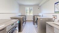 Located at 1701 Cedar Hill Cross Rd in Saanich, BC, this shared laundry facility is a thoughtfully designed, single-floor space that prioritizes both functionality and comfort. The room is finished in clean white walls and a bright textured ceiling, creating a fresh and welcoming atmosphere throughout.

At the heart of the space are two rows of stainless steel LG commercial-grade front-loading washers and dryers, arranged to maximize efficiency and ease of use. Each machine is a Direct Drive model, and usage instructions are posted prominently on the wall for resident convenience. A continuous white countertop runs along the perimeter, providing ample folding and sorting surfaces.

Natural light filters in through a frosted window at the far end of the room, offering a pleasant view of surrounding greenery. Beneath the countertop on one side, a warm wood-toned cabinetry unit houses a deep utility sink with a commercial-style spring faucet — ideal for hand-washing and pre-treating garments.

The space also includes a dedicated study and waiting area near the entrance, furnished with a long light wood communal table, bar stools, a classic Windsor chair, and a wall-mounted bookshelf stocked with reading material. Underfoot, elegant herringbone-patterned tile flooring ties the entire space together with a modern, polished aesthetic.