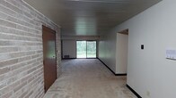 This single-floor space presents an open and functional layout centered around a generously sized main living area. The room is finished with neutral-toned carpeting that extends throughout, lending a consistent and understated feel underfoot. A distinctive exposed brick wall frames the entry, adding a rustic, textural contrast to the otherwise clean and simple interior.

The ceiling features horizontal wood paneling, contributing warmth and a mid-century character to the space. Natural light is introduced through a large sliding glass door positioned at the far end of the room, offering an unobstructed view of mature trees and lush greenery beyond — creating a peaceful, nature-connected ambiance. A floor-level vent beneath the window suggests in-floor climate control, adding to the room's practical design.

The entryway is defined by a solid brown door set within the brick wall, providing a clear transition point into the living space. An open passage on the right side of the room leads further into the unit, suggesting additional rooms beyond. The walls are painted in neutral tones, keeping the atmosphere light and versatile for a variety of uses.