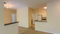 This single-floor residence offers a thoughtfully designed open-concept layout that seamlessly connects the living area to the kitchen, creating a spacious and functional flow. The kitchen is well-appointed with white cabinetry, dark countertops, a white refrigerator, and a built-in peninsula with matching dark countertop surfaces — ideal for casual dining or meal prep. The cabinetry provides ample storage, and the tile backsplash adds a clean, finished look to the cooking space. The living area features neutral wall-to-wall carpeting that contributes to a warm, inviting ambiance, while the light-toned walls reflect natural and overhead lighting to keep the space feeling bright and open. A dedicated laundry area is conveniently tucked adjacent to the main living space, housing a washer and dryer behind a classic paneled door, adding practical in-unit functionality. Additional doors suggest the presence of closets or utility storage, enhancing everyday convenience. Ceiling-mounted lighting fixtures provide consistent illumination throughout. The overall design is clean and understated, offering a versatile, move-in-ready environment with a neutral palette suited to a variety of furnishing styles.