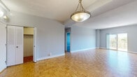 This single-floor residence offers a thoughtfully designed layout with a variety of distinct living spaces connected by open sightlines and consistent finishes throughout. The main living and dining areas are unified by warm parquet hardwood flooring arranged in a classic basket-weave pattern, complemented by soft grey walls, white baseboards, and a textured ceiling. Pendant light fixtures with brushed metal finishes anchor the dining zone, while a tray ceiling detail adds subtle architectural interest. Large sliding glass doors lead to a private balcony enclosed by ornate metal railings, offering elevated views of a lush, tree-lined landscape — particularly striking during autumn foliage. The kitchen is well-equipped, featuring rich dark wood cabinetry, granite countertops with a subtle speckled finish, a white subway tile backsplash, and stainless steel appliances including a gas range and over-the-range microwave. The bedroom is finished with plush grey carpeting, providing a quieter, softer retreat. The bathroom includes a granite-topped vanity with white cabinetry, an undermount sink, and a glass-enclosed shower, presenting a clean and functional design. Throughout, the space maintains a neutral, versatile palette suited to a range of interior styles.