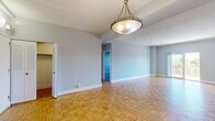 This single-floor residence offers a thoughtfully designed layout with a variety of distinct living spaces connected by open sightlines and consistent finishes throughout. The main living and dining areas are unified by warm parquet hardwood flooring arranged in a classic basket-weave pattern, complemented by soft grey walls, white baseboards, and a textured ceiling. Pendant light fixtures with brushed metal finishes anchor the dining zone, while a tray ceiling detail adds subtle architectural interest. Large sliding glass doors lead to a private balcony enclosed by ornate metal railings, offering elevated views of a lush, tree-lined landscape — particularly striking during autumn foliage. The kitchen is well-equipped, featuring rich dark wood cabinetry, granite countertops with a subtle speckled finish, a white subway tile backsplash, and stainless steel appliances including a gas range and over-the-range microwave. The bedroom is finished with plush grey carpeting, providing a quieter, softer retreat. The bathroom includes a granite-topped vanity with white cabinetry, an undermount sink, and a glass-enclosed shower, presenting a clean and functional design. Throughout, the space maintains a neutral, versatile palette suited to a range of interior styles.