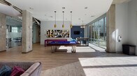 Located at 630 Cotanche St in Greenville, NC, this single-floor amenity space blends contemporary design with functional social and recreational areas. The open-plan layout is anchored by exposed concrete columns and a mix of wood-look and tile flooring, creating an industrial-modern aesthetic throughout.

The central game and lounge area serves as the heart of the space, featuring a bold blue billiards table illuminated by striking gold pendant lights. Vibrant abstract artwork adorns the walls, while a wall-mounted flat-screen TV and plush velvet seating arrangements add comfort and entertainment options. A shuffleboard table complements the game area.

Adjacent to the game zone, a relaxed lounge area is furnished with tufted sofas, accent chairs, and a round coffee table beneath a sculptural brass chandelier. A textured dark accent wall adds depth, while expansive floor-to-ceiling glass windows flood the room with natural light and offer direct views of the outdoor courtyard.

The fitness center is enclosed by full-height glass walls, providing visibility and an open feel while housing a variety of cardio and exercise equipment. A connecting corridor with track lighting links the various zones of the space.

A high-top bar area with industrial-style stools offers additional gathering space, positioned alongside the glass perimeter to maximize courtyard views. Throughout, the design balances urban sophistication with inviting, social functionality.
