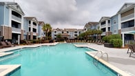 Located at 1001 Bear Island Rd in Summerville, South Carolina, this outdoor amenity space serves as a central gathering hub for the residential community. The centerpiece is a generously sized resort-style swimming pool with crystal-clear turquoise water, complemented by a separate spa or wading area accessible via a dedicated entry staircase. Surrounding the pool is a spacious sundeck outfitted with rows of lounge chairs, providing ample space for relaxation and recreation. A charming gazebo or pavilion structure anchors the far end of the pool area, offering a shaded retreat for residents. Umbrella-shaded seating areas are also scattered throughout the deck for additional comfort. Mature palm trees and manicured greenery are thoughtfully integrated throughout the space, lending a tropical, resort-like ambiance. The pool area is framed on multiple sides by the community's multi-story residential buildings, which feature a classic coastal architectural style with neutral siding, gabled rooflines, and private balconies. The overall design of this outdoor space reflects a commitment to creating a vibrant, community-centered environment in the heart of the Lowcountry.