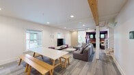This two-story communal space combines modern design with warm, inviting details across a thoughtfully arranged layout. The main floor opens into a spacious lounge area featuring large sectional sofas, a wall-mounted flat-screen TV with a media console, and built-in speakers. Exposed natural wood beams accent the white ceilings, adding warmth and architectural character. A long communal dining table with bench seating sits adjacent to the lounge, alongside a kitchenette with dark cabinetry and open shelving. A sliding barn-style door in light wood adds a contemporary touch. Intimate nook seating with corner windows floods the space with natural light and offers views of the surrounding neighborhood. The bathroom is finished with classic white subway tile, a bold purple floor, and a sleek gray countertop with trough sink. A striking staircase with light wood treads and white metal railing connects the floors. Upstairs, vaulted ceilings define several distinct areas, including a cozy lounge with modern curved chairs, a dedicated conference room with a large table and dual windows, and additional quiet seating nooks. Throughout both floors, neutral carpet with a linear pattern, recessed lighting, and a cohesive palette of white, gray, and purple accents create a clean, functional, and welcoming environment.