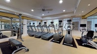 This single-floor fitness center is thoughtfully designed to accommodate a comprehensive workout experience. The space is divided into distinct functional zones, beginning with a bright, open cardio area lined with multiple treadmills and elliptical trainers, each equipped with individual display screens. Large windows fitted with plantation shutters allow generous natural light to fill the room, while a ceiling fan overhead aids in air circulation. Full-length mirrors along one wall visually expand the space and support proper form during exercise.

Adjacent to the cardio zone, a dedicated strength training area houses a selection of cable and resistance machines, including a leg press and multi-function cable systems. A neatly organized dumbbell rack holds a graduated range of free weights, accompanied by a weight bench for versatile training options.

Throughout, the décor maintains a refined, upscale character with white walls accented by gold and black trim detailing, recessed lighting, and neutral carpeted flooring. Framed artwork is displayed along the walls, adding a polished touch. Dark-framed glass double doors serve as the main entrance, contributing to the facility's clean, professional aesthetic. The overall ambiance is both functional and inviting.