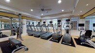This single-floor fitness center is thoughtfully designed to accommodate a comprehensive workout experience. The space is divided into distinct functional zones, beginning with a bright, open cardio area lined with multiple treadmills and elliptical trainers, each equipped with individual display screens. Large windows fitted with plantation shutters allow generous natural light to fill the room, while a ceiling fan overhead aids in air circulation. Full-length mirrors along one wall visually expand the space and support proper form during exercise.

Adjacent to the cardio zone, a dedicated strength training area houses a selection of cable and resistance machines, including a leg press and multi-function cable systems. A neatly organized dumbbell rack holds a graduated range of free weights, accompanied by a weight bench for versatile training options.

Throughout, the décor maintains a refined, upscale character with white walls accented by gold and black trim detailing, recessed lighting, and neutral carpeted flooring. Framed artwork is displayed along the walls, adding a polished touch. Dark-framed glass double doors serve as the main entrance, contributing to the facility's clean, professional aesthetic. The overall ambiance is both functional and inviting.