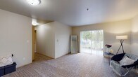 This single-floor residential unit offers a well-organized layout designed for comfortable everyday living. The living room is generously sized, featuring soft wall-to-wall carpeting and a large sliding glass door that fills the space with natural light and provides access to an outdoor area with green views. A partial dividing wall subtly separates the living room from the kitchen, which features a pass-through breakfast bar, light wood cabinetry, laminate flooring, and includes a full-size refrigerator and range. The unit includes multiple bedrooms, each finished with plush carpeting, neutral-toned walls, and ample closet space. One bedroom notably features a built-in closet with hanging rods and a window overlooking a lush outdoor setting, along with baseboard heating. The unit is served by two bathrooms: a full bath with a tub-shower combination, a single vanity, and tile flooring, and a second bath featuring a single vanity, toilet, grab bar, and matching tile floors. Connecting hallways and interior doors provide a smooth flow between all areas of the unit. The overall aesthetic is clean, neutral, and functional, making it a practical and comfortable living space.