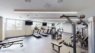 Located at 1110 W Flagler St in Miami, FL, this fitness center offers a well-equipped, modern workout facility designed to support a full range of exercise routines. The space features a clean, contemporary aesthetic with smooth light-gray walls, neutral-toned flooring, and recessed linear lighting that creates a bright and energizing atmosphere throughout.

The cardio zone is outfitted with multiple treadmills, elliptical machines, and stationary bikes, positioned along windowed walls that allow natural light to filter in and offer views of surrounding tropical greenery. Wall-mounted flat-screen televisions are positioned throughout for entertainment during workouts.

The strength training area is generously equipped with a variety of cable machines, weight benches, and selectorized resistance machines catering to both upper and lower body training. Free weights, medicine balls, and stability balls are also available, providing ample options for functional and accessory training. A large floor-to-ceiling mirror spans one wall, enhancing the sense of openness and allowing users to monitor their form.

Vibrant botanical-themed artwork adorns the walls, adding a fresh and lively touch to the space. Storage cubbies offer convenient organization for personal belongings. The facility is well-maintained, thoughtfully laid out, and designed to deliver a comprehensive fitness experience in the heart of Miami.
