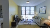 This single-floor space showcases a thoughtfully designed living area that balances modern aesthetics with a warm, inviting atmosphere. The room is anchored by a striking yellow accent wall that adds a vibrant yet soothing tone, complementing the neutral gray and beige furnishings throughout.

Large multi-pane windows with horizontal blinds dominate one wall, flooding the interior with abundant natural light while offering views of the surrounding exterior. The windows create a strong visual connection to the outdoors, enhancing the sense of openness within the space.

The living area is furnished with a plush gray sofa adorned with decorative pillows in coordinating yellow and neutral tones, paired with a sleek, chrome-framed glass coffee table that adds a contemporary touch. A minimalist white accent chair provides additional seating, while a dark wood media console houses a flat-screen television, grounding the entertainment area.

Soft, layered area rugs define the seating zone, adding texture and warmth underfoot. Overhead, a flush-mount ceiling light provides ambient illumination, supplemented by a classic table lamp in the corner. Subtle decorative elements, including small plants and ceramic accents, complete the polished, well-curated look of the space.