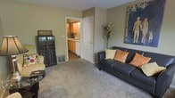 This multi-story residence spans three floors, providing ample space across a thoughtfully arranged layout. The living area serves as a central gathering space, featuring wall-to-wall carpeting that adds warmth and comfort underfoot. Neutral-toned walls create a versatile backdrop, complemented by tasteful wall art that adds character to the room. The living space flows naturally into the kitchen, which is visible through a connecting doorway and showcases warm wood-toned cabinetry, a classic tile backsplash, and white appliances. The kitchen's layout is efficient and practical, with counter space suited for everyday meal preparation. A floor vent integrated into the living area indicates a central climate system throughout the home. The overall ambiance of the main living level is casual and inviting, with a neutral color palette that lends itself to a variety of interior styles. With three full floors to explore, this residence offers distinct zones for living, sleeping, and additional functional spaces, making it a well-rounded home suited for a range of lifestyles.