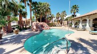 This expansive resort-style outdoor amenity space is designed for relaxation and recreation, offering a rich variety of features set against a lush backdrop of mature palm trees and desert landscaping. The centerpiece is a freeform swimming pool with crystal-clear turquoise water and a dramatic natural rock waterfall feature, evoking a true resort atmosphere. Surrounding the pool, a generous sun deck provides ample space for lounging chairs and umbrellas.

Multiple covered pavilions extend the usability of the space throughout the day. One covered area houses a fully equipped outdoor kitchen with dual large stainless steel built-in gas grills, ideal for gatherings. Nearby, a stylish open-air lounge area offers plush wicker seating with aqua-toned cushions, a fire pit table, and a striped shade sail overhead, creating a casual yet refined setting for socializing.

A separate covered colonnade-style patio, supported by elegant white columns, provides a spacious seating arrangement with neutral-toned outdoor sofas and chairs, ceiling fans overhead, and direct views of the pool. Adjacent to this is a covered recreational corridor featuring an outdoor foosball table and a shuffleboard court, lined with additional seating and ceiling fans — perfect for casual competitive play in a shaded environment.