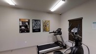 This single-floor fitness room offers a straightforward and functional workout environment designed for cardio-focused exercise. The space is finished with neutral light-colored walls and light flooring, creating a clean and bright atmosphere. Overhead fluorescent lighting ensures the room is well-illuminated, supplemented by a window fitted with dark wood blinds that allows for natural light control.

The room is equipped with a Spirit-brand treadmill and an elliptical machine, providing essential cardio options for users. A security camera is mounted near the ceiling, adding a layer of safety and monitoring to the space.

The walls are decorated with a curated selection of framed motivational posters with a boxing theme, including a bold graphic featuring the iconic Muhammad Ali quote, "Float like a butterfly, sting like a bee," alongside additional boxing-related imagery. These pieces contribute an energetic and motivating ambiance to the room.

Overall, the space is compact yet efficiently arranged, making it suitable for individual or small-group workouts. Its minimalist design keeps the focus on functionality and motivation.