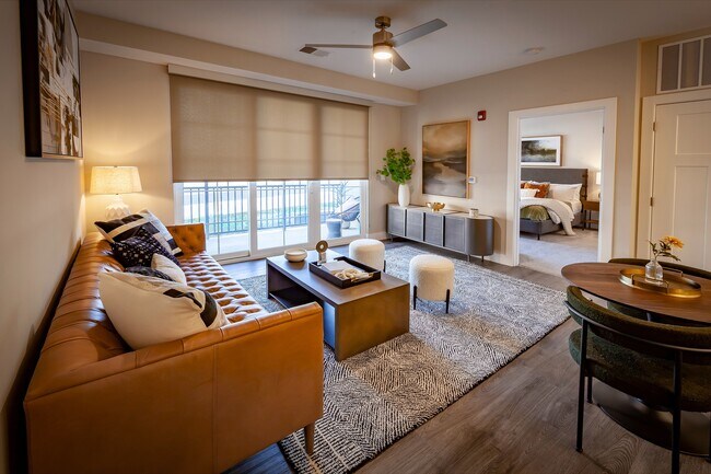 Elegant apartment living room with a chic leather sofa, modern decor, abundant natural light, and an open layout, including dining area, flowing into a cozy bedroom. - Madison at Greenwood