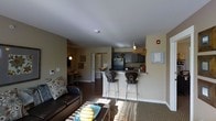 This single-story residence offers an open-concept layout that seamlessly connects the living area and kitchen, creating a cohesive and functional living space. The living room is furnished with a dark leather sofa accented by decorative throw pillows, complemented by a patterned area rug and a coffee table that anchors the seating area. Natural light filters in through unseen windows, casting warm sunlight across the floor. Dark hardwood-style flooring runs throughout the main living space, adding a rich contrast to the neutral beige and warm gray wall tones. The kitchen features dark cabinetry with a practical layout, black appliances including a full-size refrigerator, and a raised breakfast bar that provides both additional counter space and casual dining with two bar-height chairs. A doorway visible from the living area leads to additional rooms down a hallway, which includes a wall-mounted bulletin board and a pendant light fixture. Framed wall art and subtle décor accents contribute to the warm, lived-in ambiance of the space. The overall design is practical and cohesive, with consistent finishes throughout.