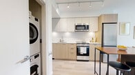 Located at 8460 Ash Street in Vancouver, BC, this thoughtfully designed one-bedroom, one-bathroom residence offers a modern and efficient layout across a single floor. The open-concept kitchen and dining area is a standout feature, showcasing two-tone cabinetry with light wood lower drawers and white upper cabinets, white quartz countertops, stainless steel appliances including a built-in microwave and range, and light hardwood-style flooring that flows throughout the unit. A compact island with a wood-top and built-in shelving provides additional dining and storage space. The living room is bathed in natural light through floor-to-ceiling sliding glass doors that open directly onto a private balcony, offering elevated views of the surrounding Vancouver cityscape and greenery. The bedroom features a large window framing the same scenic city views, creating a bright and airy retreat. The bathroom is finished with a warm wood vanity, stone countertop, and a backlit mirror, reflecting the unit's clean, contemporary aesthetic. In-unit stacked washer and dryer are conveniently tucked within a dedicated laundry closet. The entrance and additional closet complete this well-appointed, efficiently designed urban home.