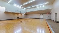This indoor sports facility is purpose-built for athletic activity, offering two distinct court areas designed to accommodate a range of sports and recreational pursuits. The centerpiece is a full-size basketball court featuring polished hardwood flooring with clearly marked red court lines and a regulation glass backboard and hoop. The court is enclosed by white-painted masonry block walls and topped with soaring ceilings that allow for unrestricted play. Rows of fluorescent strip lighting run the length of the ceiling, ensuring bright, consistent illumination throughout. A row of clerestory windows along the upper walls introduces additional natural light, enhancing the open, airy atmosphere of the space. A carpeted spectator or buffer zone lines one side of the basketball court, along with viewing windows that look into an adjacent fitness area. The facility also includes at least one dedicated racquetball court, characterized by smooth white walls, light hardwood flooring with regulation court markings, and a glass entry door for easy access. Recessed ceiling lights provide focused illumination within the racquetball enclosure. Throughout the facility, HVAC vents ensure comfortable climate control. The overall design is clean, functional, and well-maintained, making this an exceptional multi-sport recreational space.