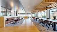 This thoughtfully designed single-floor amenity space blends contemporary aesthetics with functional versatility. The entrance lobby greets visitors with bold red accent seating, textured carpet tile flooring, a wall-mounted display, and elevator access, all framed by warm mauve accent walls and glass-paneled partitions that hint at the vibrant spaces beyond.

The main gathering area is expansive and light-filled, anchored by floor-to-ceiling windows and glass doors that open to outdoor balconies with sweeping views of the surrounding landscape and pool area. Warm hardwood floors contrast elegantly with patterned area rugs throughout. Slatted wood ceiling panels add architectural warmth and visual interest, while pendant lighting punctuates key zones.

A dedicated co-working and dining section features large communal tables with leather chairs beneath the striking wood-slat ceiling, ideal for collaborative work or group gatherings. Nearby, a lounge area with a built-in banquette, round tables, and upholstered chairs sits beneath sculptural acoustic ceiling baffles that add texture and sound management.

The bar area is a standout feature, offering a long counter with dark bar stools, pendant globe lighting, a muted blue-grey ceiling, and open shelving with curated décor. A welcoming lounge near the entrance with lush planters and bold red chairs provides an inviting transitional space, completing this well-appointed amenity floor.