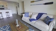 This single-floor space embraces a charming nautical aesthetic throughout its interior. The living area serves as the focal point, anchored by a classic cream-colored sofa adorned with studded detailing along its base. Coastal accents are carefully curated, including a navy blue compass-print throw pillow and a coordinating blue blanket draped casually over the couch. A pair of decorative wooden oars mounted on the wall above the sofa adds a distinctive maritime character to the space.

The flooring throughout is rich, dark hardwood-style planking, providing a warm contrast to the light-toned walls. A rustic wooden coffee table sits atop a blue-patterned area rug, further reinforcing the coastal theme. Decorative coral and nautical figurines rest on the table's surface, adding thoughtful detail to the arrangement.

A console table positioned behind the sofa provides additional surface space, while a hallway leading deeper into the unit hints at further rooms beyond. The open layout and neutral wall tones contribute to an airy, relaxed ambiance. Natural light filters through the space, enhancing the bright and breezy coastal atmosphere that defines this well-appointed interior.