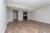 This single-floor space offers an open and functional layout characterized by a clean, neutral aesthetic. The main living area showcases a combination of rich, dark hardwood flooring and plush carpeting, creating a natural visual distinction between zones within the space. The walls are finished in a warm beige tone, lending a welcoming and versatile backdrop that suits a variety of interior styles.

A track lighting fixture is mounted on the ceiling, providing adjustable, directional illumination across the room. The ceilings feature a subtle texture and are complemented by crisp white trim and baseboards throughout, adding a polished, finished look. A white paneled door leads to an adjacent area or hallway, partially revealing a connected space beyond.

Ventilation is provided via a ceiling-mounted HVAC vent, ensuring comfortable climate control. The open wall space offers ample flexibility for furniture arrangement. Overall, the space presents a practical and well-maintained interior with a straightforward, move-in-ready appeal suited for everyday living.