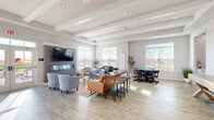 Located at 226 Bucknoll Road in Manheim, PA, this single-floor community space offers a well-appointed and thoughtfully designed layout that balances comfort with functionality. The main living area is expansive and airy, highlighted by striking exposed beam ceilings with recessed lighting that lend a clean, modern architectural character. Light wood-style flooring runs throughout, complementing the neutral gray walls and creating a warm, cohesive aesthetic. A wall-mounted fireplace with a sleek tile surround anchors one side of the living area, flanked by a large flat-screen television and comfortable seating arrangements. Generous windows along multiple walls flood the space with abundant natural light and offer pleasant outdoor views of the surrounding landscape. A dedicated game table area provides an inviting spot for recreation, adding versatility to the room. Adjacent high-top bar tables offer additional casual seating. The kitchen is crisp and functional, featuring white shaker cabinetry with matte black hardware, light quartz countertops, stainless steel appliances, and its own natural light source. A connected entryway with glass double doors creates a welcoming transition into the space. Overall, the environment is polished, bright, and well-suited for gathering and community use.