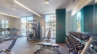 Located on Delaware Street in Denver's 80204 neighborhood, this fitness center is a fully outfitted workout facility designed to support a wide range of exercise routines. The space is laid out across a single floor with a practical, open floor plan that maximizes accessibility to equipment throughout.

The cardio zone features a row of Matrix treadmills and elliptical machines positioned along large floor-to-ceiling windows, flooding the area with natural light and offering views of the surrounding streetscape. A bank of full-length mirrors lines one wall, visually expanding the space and allowing users to monitor their form.

The strength training section is well-stocked with a comprehensive rack of dumbbells arranged along a mirrored wall, multiple Matrix cable and functional training machines, adjustable benches, and a variety of cable pulley systems suited for both upper and lower body workouts. Additional specialty machines for rowing and targeted muscle training are distributed throughout the floor.

Underfoot, durable patterned carpet tiles in a warm brown tone provide both comfort and traction. Overhead, recessed and linear LED lighting supplements the abundant natural light from the large windows. A teal accent column adds a modern architectural touch to the interior design. The overall ambiance is clean, modern, and professional, making it a functional and inviting environment for daily fitness use.
