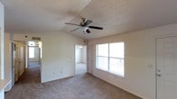 This single-floor space presents a straightforward and functional layout. The main room is finished with wall-to-wall carpeting in a neutral tone, contributing to a comfortable, understated atmosphere. A textured popcorn ceiling adds a classic touch overhead, while a multi-blade ceiling fan with an integrated light fixture provides both airflow and ambient lighting. A window fitted with horizontal blinds allows natural light to filter into the space, lending a bright quality to the room.

An open doorway connects the main room to an adjacent space, which also features carpeting and its own ceiling fan with a light fixture, suggesting it may serve as an additional living area or bedroom. A closed door nearby provides access to a separate room or closet, adding to the overall functionality of the layout. The walls throughout are painted in a light, neutral tone, creating a clean and versatile backdrop. Electrical outlets and light switches are positioned at standard heights along the walls. Overall, the space offers a practical, move-in-ready environment with modest finishes suited for everyday living.
