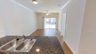 This single-floor space offers a clean, open layout that seamlessly connects the kitchen and living areas. The kitchen is appointed with rich, dark granite countertops and a stainless steel double-basin sink, providing a practical and stylish workspace. Crown molding runs throughout the space, adding a refined architectural detail to the neutral, warm-toned walls.

The living area is finished with smooth hardwood-style flooring that extends throughout, creating a cohesive and inviting atmosphere. A ceiling fan is centrally positioned overhead, promoting airflow and comfort. Natural light pours generously into the space through a large sliding glass door, which opens to an outdoor balcony, offering views and a seamless connection to the exterior.

Crown molding accents the transition between the walls and ceiling, lending a polished, well-finished feel to the overall interior. The space benefits from a bright, airy ambiance thanks to its light-colored walls and the abundance of natural light from the balcony access. The layout is versatile and well-proportioned, making it well-suited for comfortable everyday living.