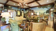 This refined single-floor space exudes classic charm and thoughtful design at every turn. The main lounge area immediately draws the eye upward to its striking coffered ceiling, defined by rich, dark wood beams that add architectural depth and warmth. A grand multi-arm chandelier hangs as a centerpiece, casting a welcoming glow across the room. Dominating the walls is an expansive hand-painted pastoral mural depicting serene countryside landscapes with trees, open fields, and winding paths — a distinctive artistic feature that lends the space a timeless, gallery-like quality. The furnishings are carefully curated to match the traditional aesthetic, including plush upholstered sofas and armchairs in soft sage and green tones, complemented by woven wicker accent chairs and dark wood coffee and side tables. An area rug anchors the central seating arrangement, creating an intimate yet open gathering space. Recessed lighting supplements the chandelier, ensuring the room is well-lit throughout. Adjacent areas visible through the elegant wood-trimmed doorways suggest additional seating and dining spaces, hinting at the broader functionality of this inviting environment. Overall, the space strikes a harmonious balance between refined elegance and comfortable livability.
