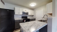 Located at 1919 Georgesville Square in Galloway, Ohio, this single-floor residence offers a well-designed layout with contemporary finishes throughout. The kitchen is a standout feature, showcasing a two-tone cabinet design with crisp white upper cabinets and dark lower cabinets, all complemented by granite-style countertops. Stainless steel appliances, including an over-the-range microwave, are paired with a black refrigerator and range. A peninsula provides additional counter space and separates the kitchen from the open living area, which flows seamlessly and features wood-look plank flooring in a neutral gray tone. The galley-style kitchen extension offers generous cabinet and counter space on both sides, along with a built-in dishwasher. The living and bedroom areas are fitted with ceiling fans and receive natural light through blinds-equipped windows. Bedrooms feature the same wood-look flooring and neutral gray walls, creating a clean, cohesive aesthetic. The bathroom includes a white vanity with ample storage, a tub-shower combination, and coordinating wood-look flooring. Throughout the unit, consistent neutral tones, modern hardware, and thoughtful storage solutions contribute to a practical and polished living environment.
